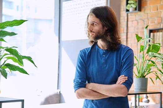 Portrait of young serious confident handsome man with long hair and beard, with crossed arms, profile view, copy space