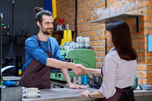 Young man in apron shaking hands with woman owner of coffee shop cafeteria restaurant, colleagues partners working together. Cooperation, small business, partnership, teamwork team, staff, success