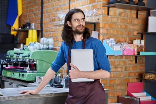 Young man in apron, food service worker, small business owner entrepreneur holding laptop near counter of coffee shop cafe cafeteria. Staff, occupation, successful business, work