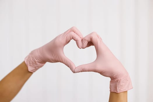 Person in pink latex gloves showing heart gesture against white background, closeup on hands. Hands with gloves making heart shape symbolize love or support to medical team.