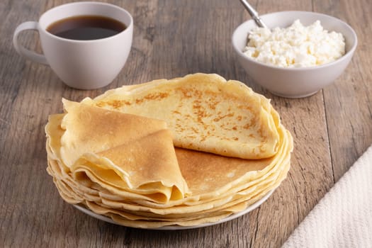 A stack of thin pancakes, a plate with cottage cheese and coffee on a wooden table.