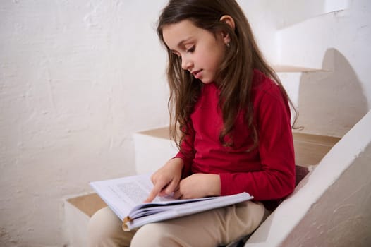 Caucasian cute child, elementary age smart little kid girl leafing through the pages of a hardcover book, reading a fairytale, sitting on the steps at home, over white wall background of a rural house