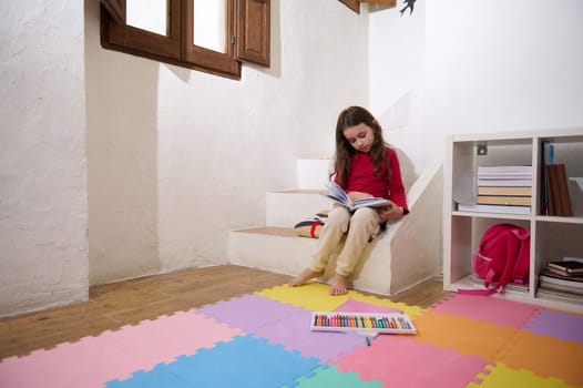 Smart elementary age girl leafing through the pages of a book, reading a fairytale, sitting on the steps barefoot on multicolored puzzle carpet at home, over white wall background of a rural house.