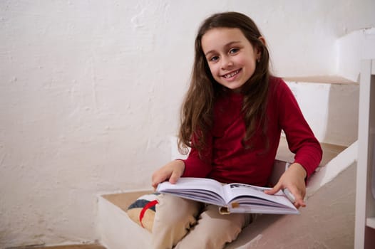 Authentic portrait of an adorable child girl holding a book, smiling looking at camera, sitting on steps at home over white wall background. Kids education and entertainment. Back to school