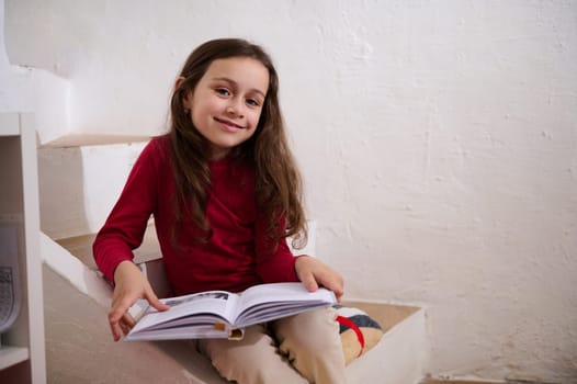 Smart elementary school student girl smiles at camera, sitting on steps at home with a book in hands. Cute child reading book. World Book Day concept. Back to school on new semester of academic year