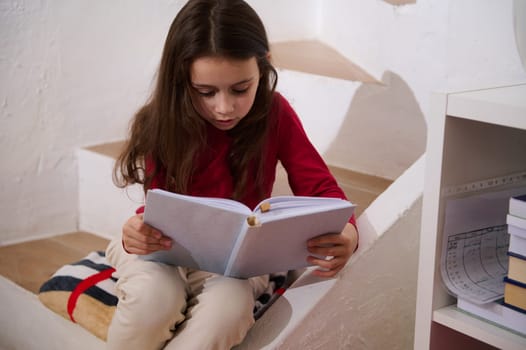 World Book Day. Portrait of cute little school aged girl focused, holding book and reading text, sitting on steps at cozy home interior. Copy space for advertising text