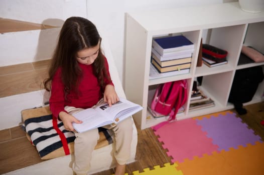 View from above school age child girl reading a book, sitting on steps of her room with multicolored puzzle carpet. The concept of back to school. Smart kids. Erudition and learning. World Book's Day.