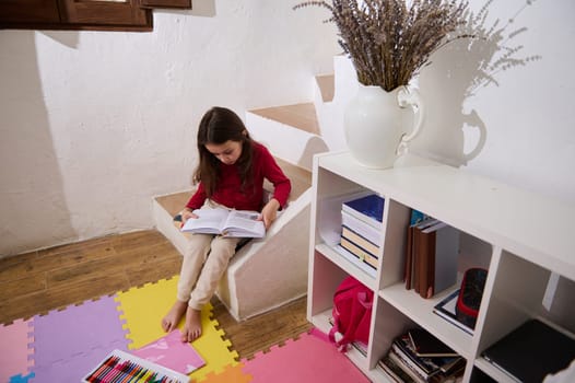 View from above on erudite child girl, elementary age schoolgirl reading a book in her room. Nearby bookshelf with books and school supplies. multi colored puzzle carpet on the floor. Autism concept