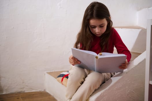 Caucasian cute little girl sitting on steps at home, reading a book over white wall background. Literature. Erudition. Knowledge. World Book Day. Back to school concept. Copy advertising space