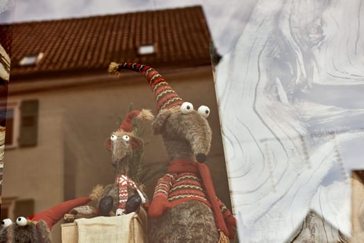 Whimsical New Year's Mouse in Cap and Scarf Stands Behind Store Display. Capture the charm of the holiday season with this delightful image featuring a toy mouse or rat wearing a comical New Year's cap and scarf, playfully standing behind a shop window display. The festive scene adds a touch of whimsy to your creative projects, invoking the spirit of celebration and joy.