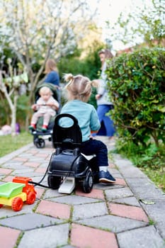Little girl drives a toy car along a path in the garden with a toy truck tied to the seat. Back view. High quality photo