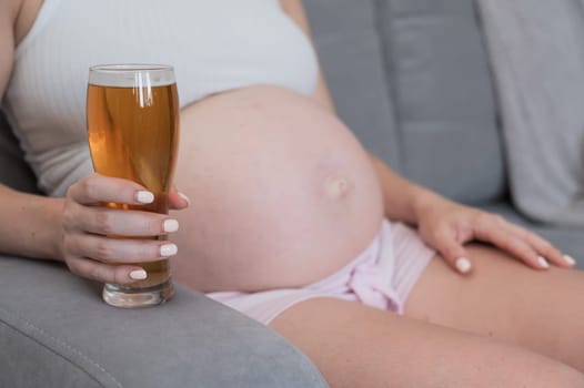Close-up of the belly of a pregnant woman holding a glass of beer while sitting on the sofa