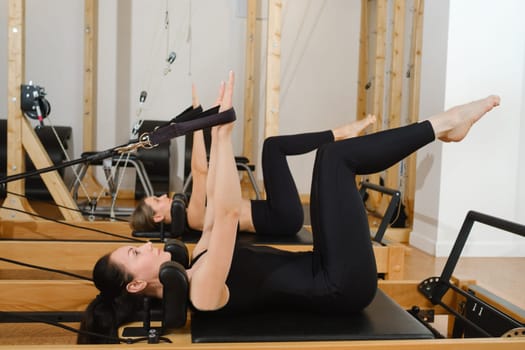 Two women are practicing physical fitness exercises on a Pilates machine in a room with hardwood flooring. Their movements showcase a beautiful balance of art and exercise