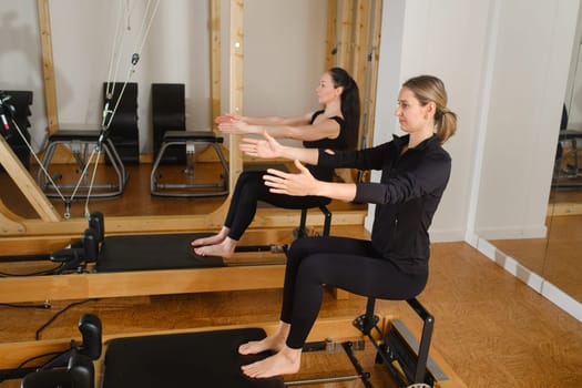 Two women are practicing physical fitness exercises on a Pilates machine in a room with hardwood flooring. Their movements showcase a beautiful balance of art and exercise