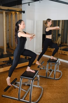 Two girls doing Pilates on a Pilates chair in a Pilates gym. A beautiful slim woman in black clothes performs exercises to strengthen the spine and leg muscles.