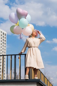 A woman with colored hair with an armful of balloons against a blue sky. Vertical photo