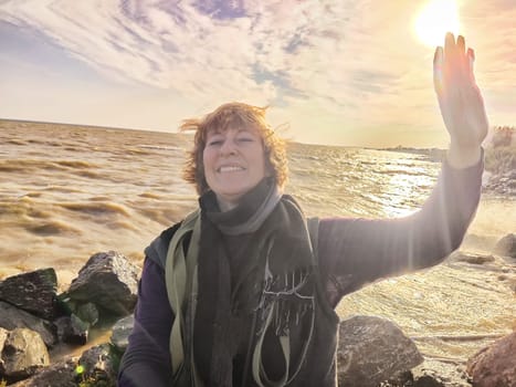 Happy cheerful middle aged woman in scarf taking selfie on nature outdoors and sun with water and waves of sea on the background