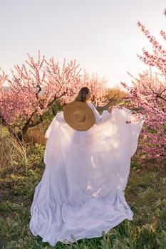 Woman blooming peach orchard. Against the backdrop of a picturesque peach orchard, a woman in a long white dress and hat enjoys a peaceful walk in the park, surrounded by the beauty of nature