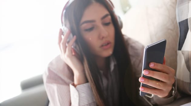Young beautiful woman headphones holds smartphone in hand using at home costs about sofa. Verifies internal information its financial advertising freedom makes money level transfer contractor concept