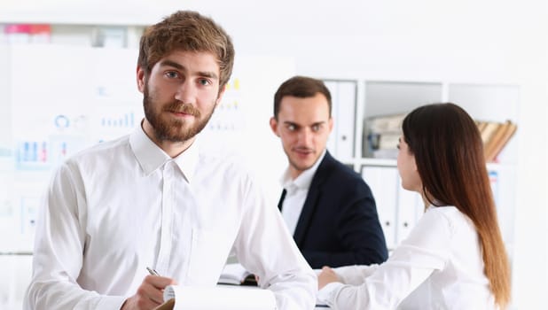 Handsome smiling bearded man in office look in camera with colleagues in background. White collar worker modern lifestyle graduate college study report idea coach train visit make note concept