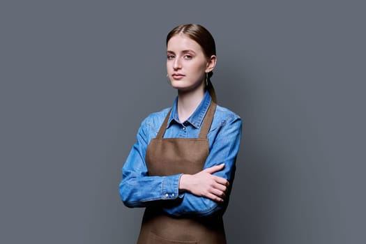 Portrait of young serious confident woman in apron on grey studio background. Successful positive female with crossed arms. Worker, startup, small business, service sector, staff, youth concept