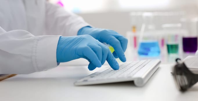 Employee chemical laboratory teacher chemist knocks his fingers keyboard makes notes electronic journal records the analysis data from reactions examining test tubes with the substance arm in gloves.