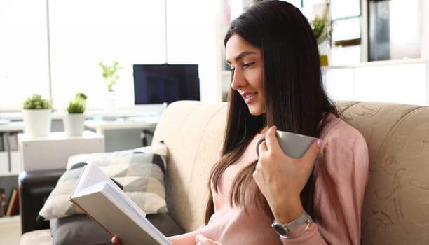 Young attractive indian teenager girl look into open book drink tea lying at couch portrait. Beautiful woman on sofa use datebook synopsis exam preparation delivery concept