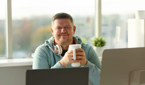 Handsome smiling male student using online education service and drink tea or coffee. Young man looking in laptop display watching training course and listening it with headphones.
