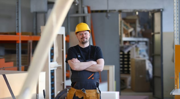 Portrait of young attractive man in work clothes and yellow helmet crossed arms smiling at shop for manufacturing furniture and details home interior