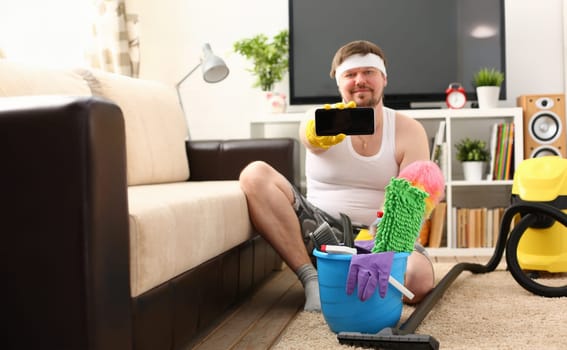 A young smiling man holds a smartphone in his hand demonstrating it to the camera advertising a mobile application to search for cleaning