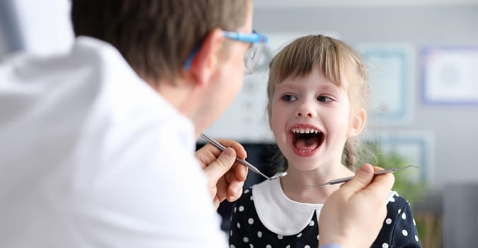 Male dentist look at open mouth litle happy girl child againlt hospital office background portrait. Tooth treatment concept