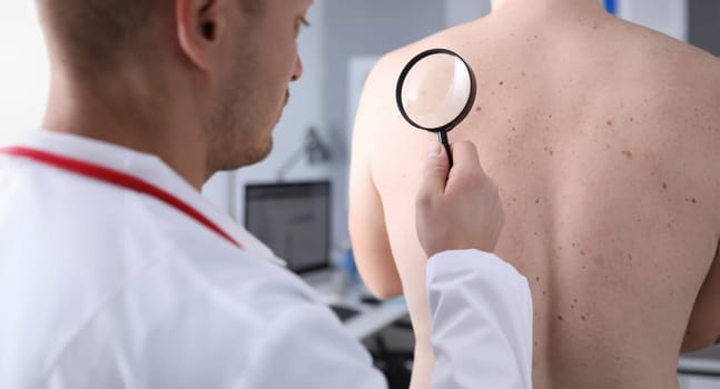 Portrait of doctor with stethoscope examining mole of males back with magnifying glass in medical office. Dermatology consultation check-up and medicine concept