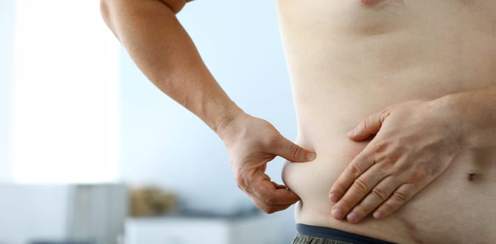 Close-up of male hand measuring thick waist to track down rate of getting plump and looking at overweight belly. Fat man preparing for training. Severe obesity and losing weight concept