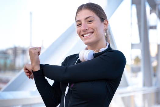 Young happy sportswoman in sports headphones doing stretching smiling looking at camera outdoors.