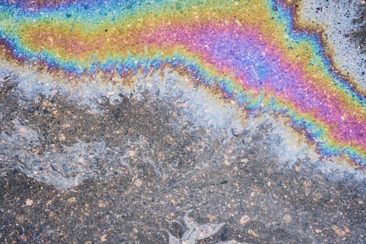 Close-up of an iridescent oil or gasoline spill on a wet asphalt, viewed from above. Bold multicolored spots on the asphalt.