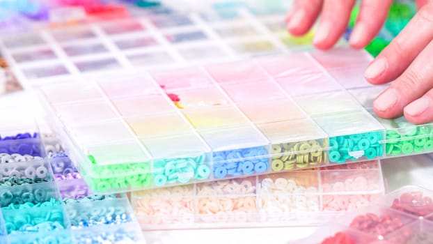 Woman hands gracefully poised over a collection of beads, sorted by color in transparent organizers. The array of beads spans a vibrant spectrum, from deep purples to bright oranges, meticulously arranged for easy selection as she embarks on creating a custom piece of jewelry.