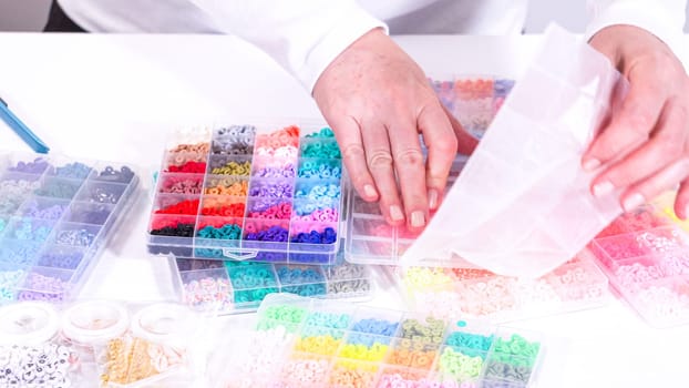 Woman hands gracefully poised over a collection of beads, sorted by color in transparent organizers. The array of beads spans a vibrant spectrum, from deep purples to bright oranges, meticulously arranged for easy selection as she embarks on creating a custom piece of jewelry.