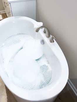 Elegant white bathtub filled with water, featuring a sleek brushed nickel faucet with water flowing gently into the tub. The bathtub modern design and the clear, tranquil water suggest a peaceful bathroom setting.