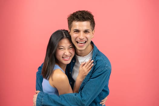 Loving young man hugging his pretty girlfriend on pink studio background, happy multiracial lovers enjoying time together, hugs, copy space.