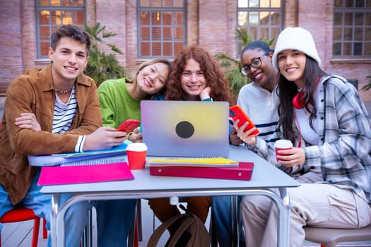 A multiethnic group of five students with a computer and notes sit near campus to review the exam Looking at camera outdoors.