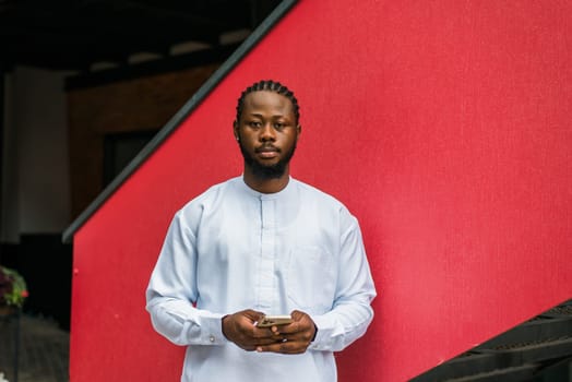 Red banner portrait of a happy african american man smiling wearing ethnic dashiki clothes outdoor in summer street wall and holds smartphone with social networks. Millennial generation student and youth. Copy space.