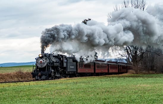 Ronks, Pennsylvania, USA, December 30, 2023 - A steam train is traveling down the tracks, leaving a trail of smoke behind it. The train is surrounded by a lush green field, creating a serene and peaceful atmosphere