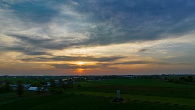 Aerial View of Dramatic Sunset Casting A Warm Glow Over A Rural Landscape With Silhouetted Trees And Buildings Under A Cloud-Streaked Sky.