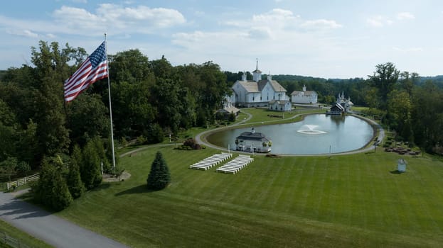 Aerial View Of A Grand White Building With A Grey Roof Surrounded By Lush Green Lawns, A Large Reflective Pond With Fountains, And A Majestic Flagpole Bearing The American Flag.