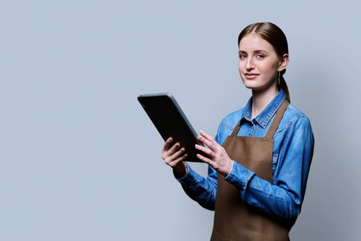 Young smiling female worker in apron uniform holding digital tablet on gray studio background. Digital technologies in business work, Internet apps applications for online orders and customer service