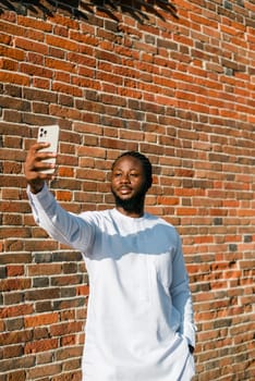 Happy young African American man in dashiki ethnic clothes taking selfie on brick wall background. Millennial generation student and youth