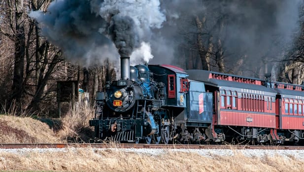 Ronks, Pennsylvania, USA, February 17, 2024 - A steam train is traveling down the tracks, leaving a trail of smoke in its wake. The train is surrounded by trees, and the smoke billows out of the engine, creating a sense of movement and power