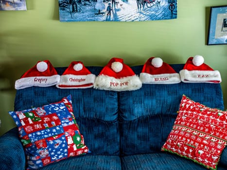 A group of five red santa hats are sitting on a blue couch. The hats are personalized with names and are arranged in a row. The couch is decorated with a red and white pillow and a blue pillow