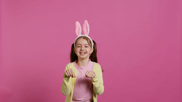 Joyful carefree schoolgirl jumping around in studio, imitating a rabbit and hopping against pink background. Cheerful active child wearing bunny ears and bouncing, adorable kid. Camera B.