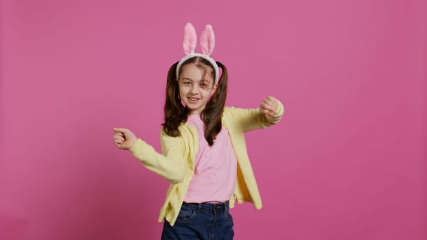 Confident cheery girl showing dance moves in the studio, feeling cheerful and positive about easter holiday festivity. Talented child dancing around and wearing bunny ears. Camera B.